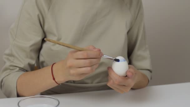 Eine unkenntliche Frau bemalt Ostereier mit einem Pinsel. Auf dem Tisch liegt ein Glas Wasser und eine Schüssel mit Eiern. Vorbereitung auf die Feiertage. — Stockvideo