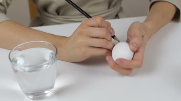 Eine unkenntliche Frau zeichnet eine Bleistiftskizze auf ein Osterei. Auf dem Tisch liegt ein Glas Wasser und eine Schüssel mit Eiern. Vorbereitung auf die Feiertage. — Stockvideo