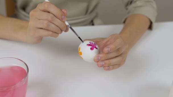 Oigenkännlig kvinna målar påskägg med en borste. På bordet är ett glas vatten och en skål med ägg. Förbereda för semester. — Stockvideo