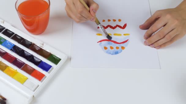 Eine unkenntliche Frau malt eine Skizze mit leuchtenden Farben auf Papier für eine Osterkarte. Auf dem Tisch liegt eine Palette mit Farben und einem Glas Wasser. — Stockvideo
