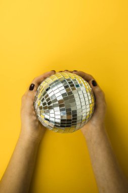 Kadın elleri, parlak sarı arka planda disko aynalı top şeklinde Noel oyuncağını düzgünce tutuyor. Yukarıdan bak. Düz yatıyordu. Mesaj için yer. Kopyalama alanı.