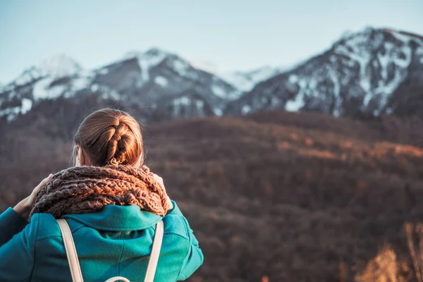 Young girl in turquoise coat with backpack enjoys walk through the beautiful autumn yellow and orange forests and views of the snow-capped mountains. Tourist route. Trees in colorful foliage.