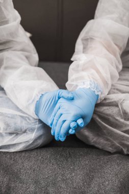 Koruyucu beyaz yarı şeffaf takım elbiseli, mavi lastik eldivenli, tıbbi maskeli iki kişi evdeki kanepede oturuyor. Koronavirüs salgını sırasında evde kal..