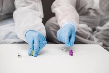 Koruyucu beyaz yarı şeffaf takım elbiseli, mavi lastik eldivenli, tıbbi maskeli iki kişi evdeki kanepede oturup masa oyunları oynayıp zar atıyorlar. Coronavirus salgını sırasında evde kal..