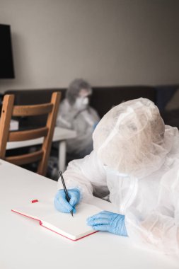Koruyucu beyaz yarı şeffaf takım elbiseli, mavi lastik eldivenli, tıbbi maskeli bir kız evde oturuyor ve çizim defterine kalemle resim çiziyor. Koronavirüs salgını sırasında tasarımcı ve sanatçının uzaktan çalışması.