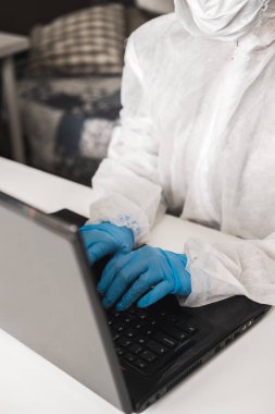Koruyucu beyaz yarı şeffaf takım elbiseli, mavi lastik eldivenli, tıbbi maskeli evde oturuyor ve karantina sırasında dizüstü bilgisayarda çalışıyor ya da çalışıyor. Koronavirüs salgını sırasında uzaktan çalışma.