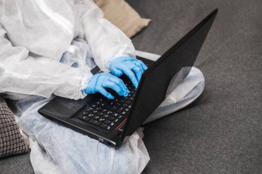 Koruyucu beyaz yarı şeffaf takım elbiseli, mavi lastik eldivenli, tıbbi maskeli evde oturuyor ve karantina sırasında dizüstü bilgisayarda çalışıyor ya da çalışıyor. Koronavirüs salgını sırasında uzaktan çalışma.