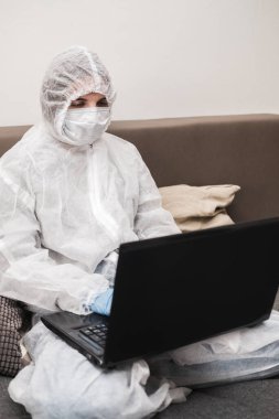 Koruyucu beyaz yarı şeffaf takım elbiseli, mavi lastik eldivenli, tıbbi maskeli evde oturuyor ve karantina sırasında dizüstü bilgisayarda çalışıyor ya da çalışıyor. Koronavirüs salgını sırasında uzaktan çalışma.