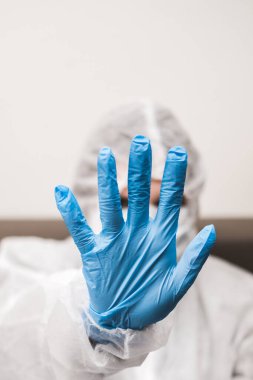 Koruyucu beyaz yarı şeffaf takım elbiseli, mavi lastik eldivenli, tıbbi maskeli biri elini öne uzatıyor. Coronavirüs salgını sırasında el kol hareketleri evde kalır..