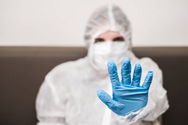Koruyucu beyaz yarı şeffaf takım elbiseli, mavi lastik eldivenli, tıbbi maskeli biri elini öne uzatıyor. Coronavirüs salgını sırasında el kol hareketleri evde kalır..