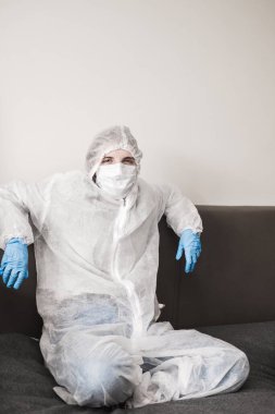Koruyucu beyaz yarı şeffaf takım elbiseli, mavi lastik eldivenli, tıbbi maskeli iki kişi evdeki kanepede oturuyor. Koronavirüs salgını sırasında evde kal..