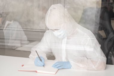 Koruyucu beyaz yarı şeffaf takım elbiseli, mavi lastik eldivenli, tıbbi maskeli bir kız evde oturuyor ve çizim defterine kalemle resim çiziyor. Koronavirüs salgını sırasında tasarımcı ve sanatçının uzaktan çalışması.