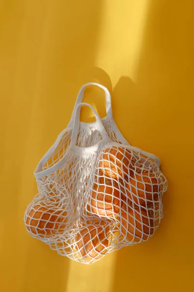 Oranges lie in string bag on bright yellow background and hard sunshine. The concept of zero waste. Using reusable bags instead of disposable cellophane packets.