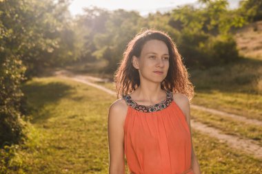 Koyu kıvırcık saçlı, parlak turuncu elbiseli güzel bir kız sonbaharda, yaz ormanlarında ya da gün batımında tarlada yürüyor. Kadın gülümsüyor ve mutlu. Romantik bir ruh hali. Neşe.