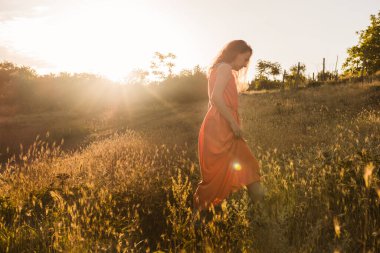 Koyu kıvırcık saçlı, parlak turuncu elbiseli güzel bir kız gün batımında bir sonbahar ormanında ya da tarlada yürüyor. Kadın gülümsüyor ve mutlu. Romantik ruh hali.