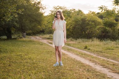 Kahverengi saçlı, beyaz elbiseli güzel bir kadın elinde akıllı telefonuyla sonbahar ya da yaz ormanlarında yürüyor. Kız ormanda mobil iletişim ve internet arıyor..