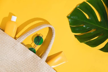 Hasır hasır torba, güneş kremi, yeşil güneş gözlüğü ve canavar yaprağıyla renkli arka planda parlak sarı düz bir döşeme. Sert gölgeler ve ışık. Üst Manzara. Kopyalama alanı.