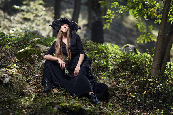 Model girl in a black dress and black hat in a mystical forest. Mystery, beauty, style, halloween portrait.