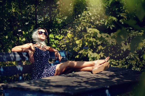 Modelo menina em um chapéu e óculos de sol sentado em um banco no par — Fotografia de Stock