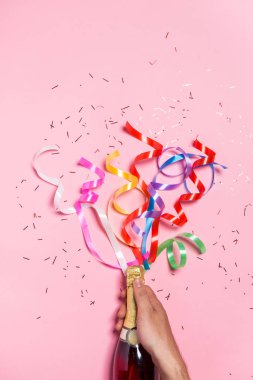 Kutlamanın düzleşmesi. Pembe arka planda renkli parti flamaları olan şampanya şişesi..