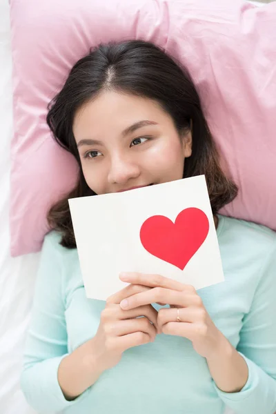 Fiatal ázsiai nő ágyon, holding üdvözlés kártya-val piros h — Stock Fotó