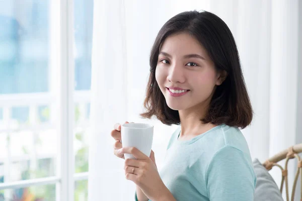 Young pretty woman sitting at opened window drinking coffee and — Stock Photo, Image