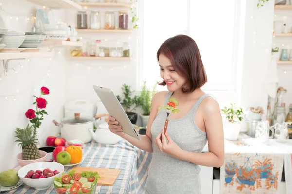 Feliz hermosa mujer asiática preparando comida saludable en la cocina y leyendo la receta usando tableta digital . —  Fotos de Stock