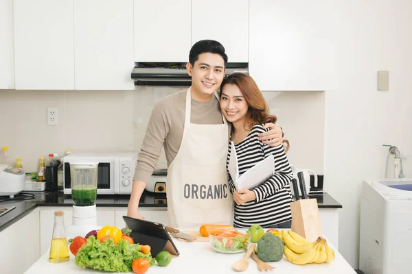 Happy asian couple following recipe on digital tablet when cooki