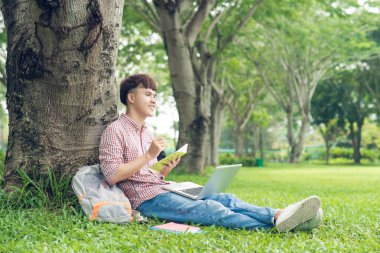 Gülümseyen rahat öğrenci uzak kampüsünde seyir çimenlerin üzerinde oturan bir