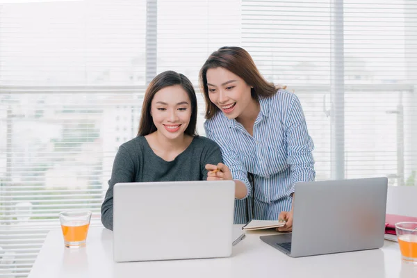 Teamwork concept with two smiling young business women sitting in office at table and working with laptops