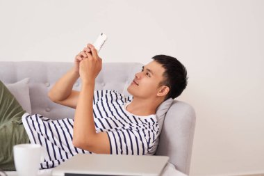 Evde oturma odasında kanepede yatan bir akıllı telefon kullanan rahat Asyalı adam