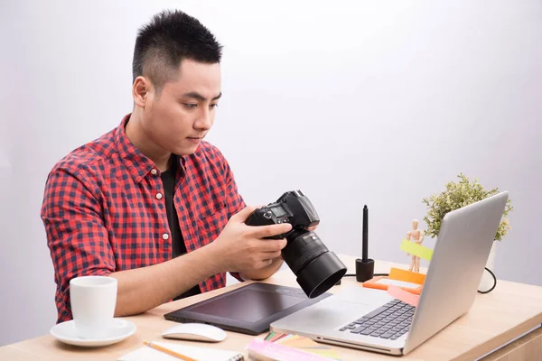 Professional photographer. Portrait of confident young man in shirt holding hand on camera while sitting at his desk.