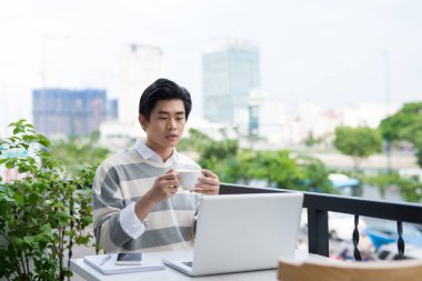 Dizüstü bilgisayarda çalışan ve kafede kahve tadını çıkarırken gülümseyen yakışıklı Asya genç adam