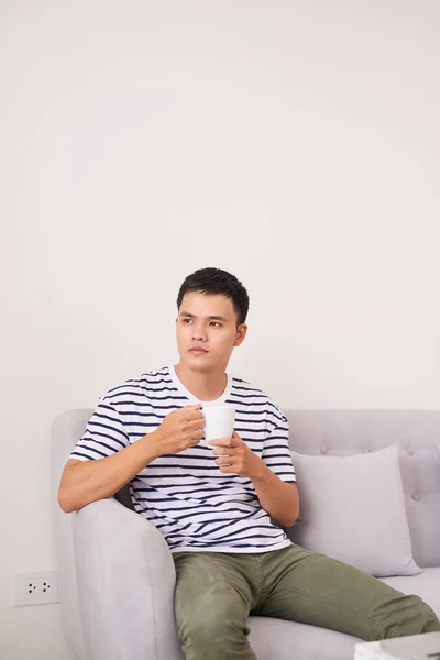 Νεαρός Ασίας Που Κάθεται Στον Καναπέ Πίνοντας Ένα Φλιτζάνι Του — Φωτογραφία Αρχείου