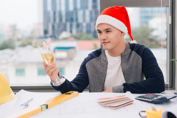 Masculino Ásia Arquiteto Santa Chapéu Sentado Mesa Segurando Copo Champanhe — Fotografia de Stock