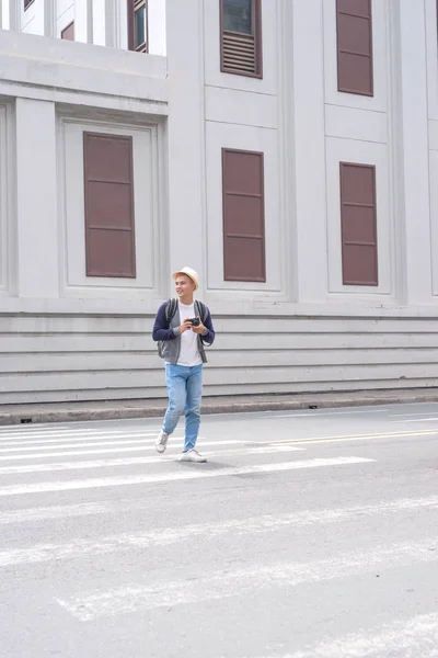 年轻的亚裔男子在帽子行走使用智能手机拍照城市 — 图库照片