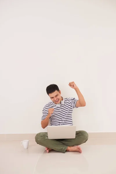 Young man playing on laptop sitting on floor enjoying win or goo — Stock Photo, Image