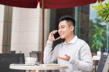 Yakışıklı genç adam içme kahve Cafe masada oturan ve Smartphone'da konuşuyor 