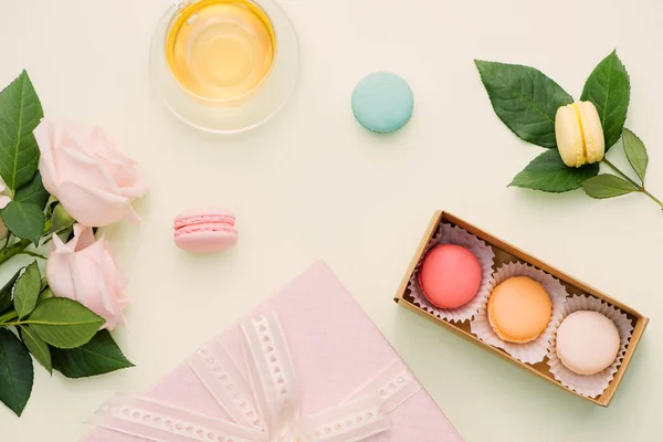 Colorful macaroons in gift box and roses with tea on light table. Top view