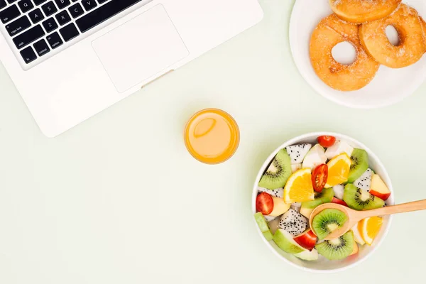 Healthy business lunch in office, fruit salad bowl near laptop on white background. Organic meal and donuts. Snack at break time.