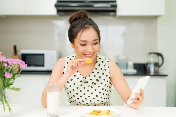Young smiling woman eating cookies and smiling, using smartphone. Healthy breakfast.