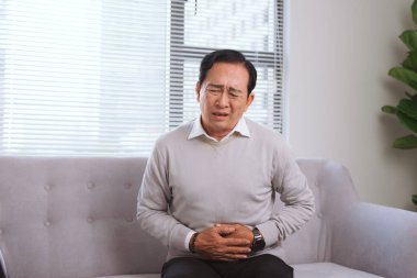 Senior asian man with stomach ache sitting on couch  clipart