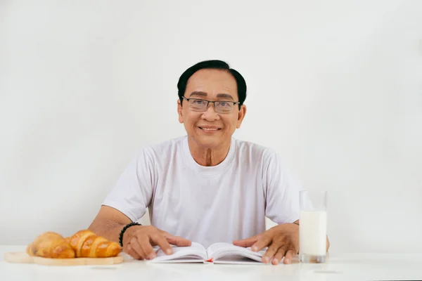 Lycklig Senior Man Äter Croissant Och Mjölk Frukost Hemma Läsa — Stockfoto