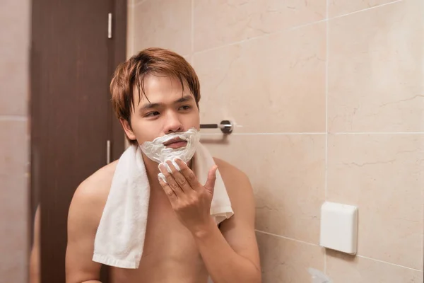 Handsome young asian man shaving his face and looking at the mirror.