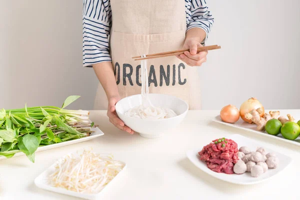 Female chef cooking  traditional Vietnamese soup Pho bo with vegetables, herbs, meat, rice noodles