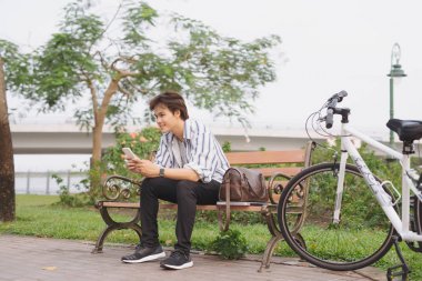 Genç adam parkta bisikletle bankta otururken telefonu kullanıyor.