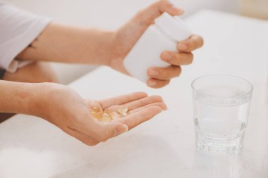 hands with yellow capsules of omega 3, white pills of glucosamine and calcium in plastic box, glass with water at wooden table, top view. clipart