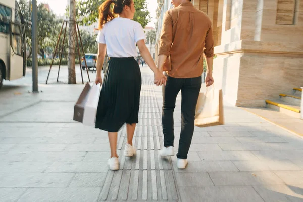 Lower body section of a young tourist couple walking by store windows and holding paper shopping bags in a destination city.