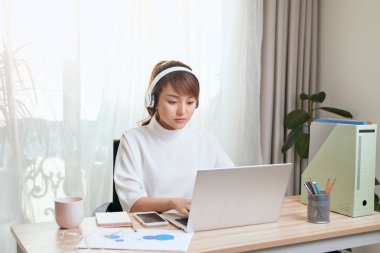 Genç Asyalı iş kadını müzik dinliyor ve evde dizüstü bilgisayarla çalışıyor.