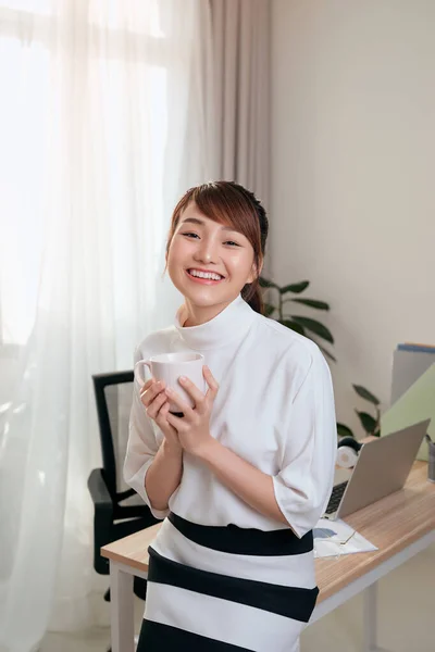 若いアジアの実業家は彼女のオフィスでコーヒーカップを保持しながら机の上に座って — ストック写真